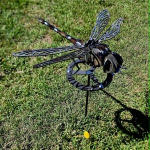 Pode incluir: Uma escultura de lib&eacute;lula em metal escuro com padr&otilde;es de asas vazados e intrincados e um corpo segmentado. A lib&eacute;lula est&aacute; montada numa base circular de metal com uma estaca vertical, criando uma pe&ccedil;a de adorno detalhada para o jardim.