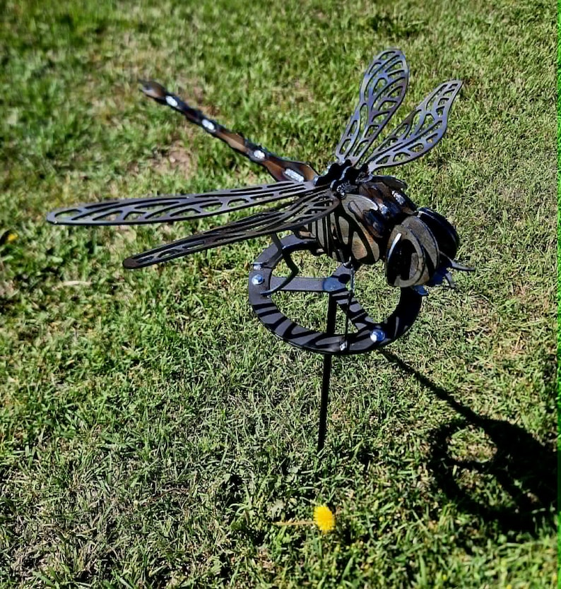 Pode incluir: Uma escultura de lib&eacute;lula em metal escuro com padr&otilde;es de asas vazados e intrincados e um corpo segmentado. A lib&eacute;lula est&aacute; montada numa base circular de metal com uma estaca vertical, criando uma pe&ccedil;a de adorno detalhada para o jardim.