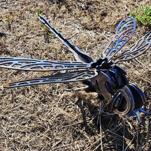 Pode incluir: Uma escultura de lib&eacute;lula em metal com padr&otilde;es intrincados de asas vazadas e um corpo segmentado. A pe&ccedil;a apresenta um acabamento escuro tratado termicamente com tons azuis e roxos iridescentes vis&iacute;veis nas asas e na cabe&ccedil;a, feita de a&ccedil;o soldado.