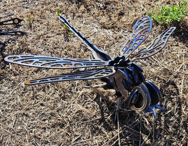 Pode incluir: Uma escultura de lib&eacute;lula em metal com padr&otilde;es intrincados de asas vazadas e um corpo segmentado. A pe&ccedil;a apresenta um acabamento escuro tratado termicamente com tons azuis e roxos iridescentes vis&iacute;veis nas asas e na cabe&ccedil;a, feita de a&ccedil;o soldado.