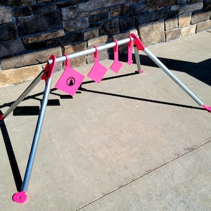 May include: A pink metal shooting target stand with five pink targets. The stand is made of metal tubing and has a triangular base. The targets are attached to the stand with metal hooks.
