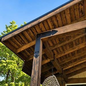 May include: A wooden porch roof with exposed beams and a black metal corner bracket. The roof is made of stained wood planks. A hanging wicker chair is suspended from the ceiling. The sky is blue and a tree is visible in the background.