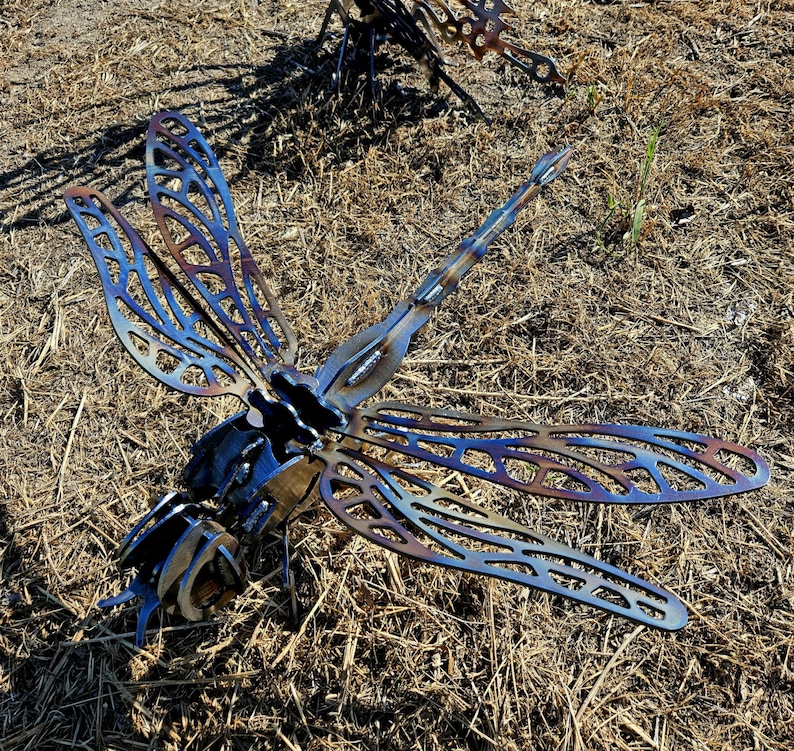 Pode incluir: Uma escultura de lib&eacute;lula em metal com padr&otilde;es de asas intrincados cortados a laser e acabamentos tratados termicamente em tons de azul iridescente e bronze. O corpo &eacute; constru&iacute;do com componentes de a&ccedil;o soldados com uma cauda segmentada.