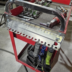 Puede incluir: Un carro de herramientas de metal rojo con un asa plateada y un estante para herramientas plateado. El carro contiene varias herramientas, incluyendo llaves de vaso, trinquetes y herramientas eléctricas. El carro está diseñado para organizar y transportar herramientas.