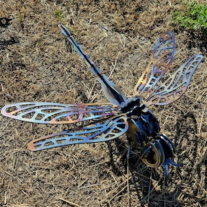 Pode incluir: Uma escultura de lib&eacute;lula em metal feita de componentes de a&ccedil;o soldado. As asas apresentam padr&otilde;es recortados intrincados com um acabamento iridescente tratado termicamente, exibindo tons de azul, roxo e dourado.