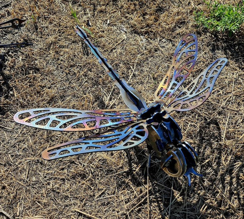 Pode incluir: Uma escultura de lib&eacute;lula em metal feita de componentes de a&ccedil;o soldado. As asas apresentam padr&otilde;es recortados intrincados com um acabamento iridescente tratado termicamente, exibindo tons de azul, roxo e dourado.