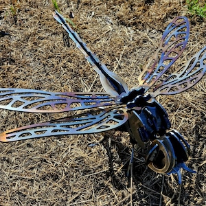 Pode incluir: Uma escultura de lib&eacute;lula em metal com padr&otilde;es de asas intrincados e vazados em tons iridescentes de azul e bronze tratados termicamente. O corpo &eacute; constru&iacute;do com componentes de a&ccedil;o em camadas, exibindo uma cauda segmentada e olhos grandes e redondos.