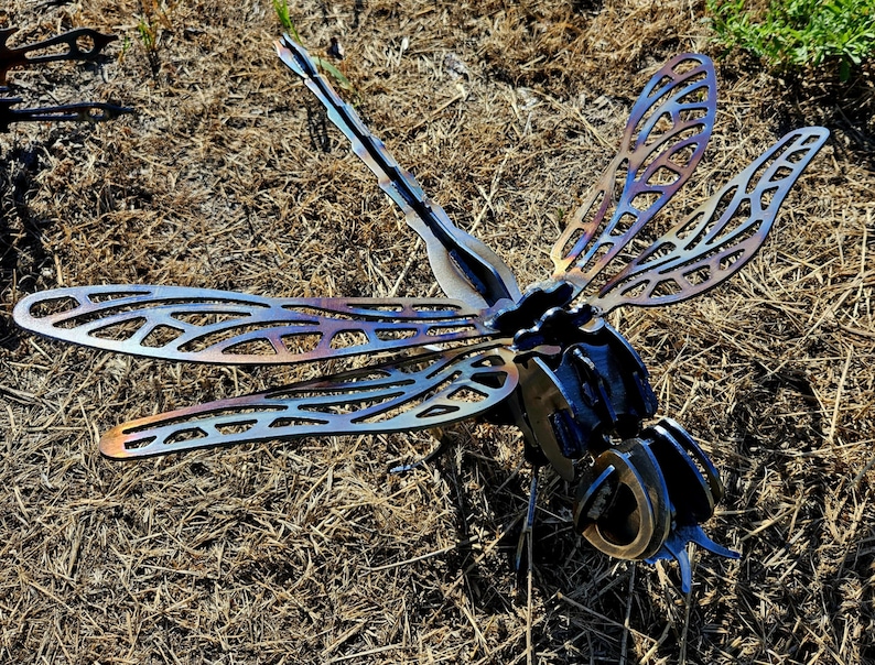 Pode incluir: Uma escultura de lib&eacute;lula em metal com padr&otilde;es de asas intrincados e vazados em tons iridescentes de azul e bronze tratados termicamente. O corpo &eacute; constru&iacute;do com componentes de a&ccedil;o em camadas, exibindo uma cauda segmentada e olhos grandes e redondos.