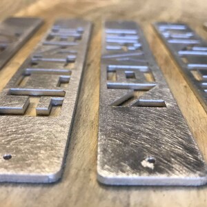 May include: Five silver metal rectangular signs with a geometric design cut out. The signs are arranged in a row on a wooden surface.