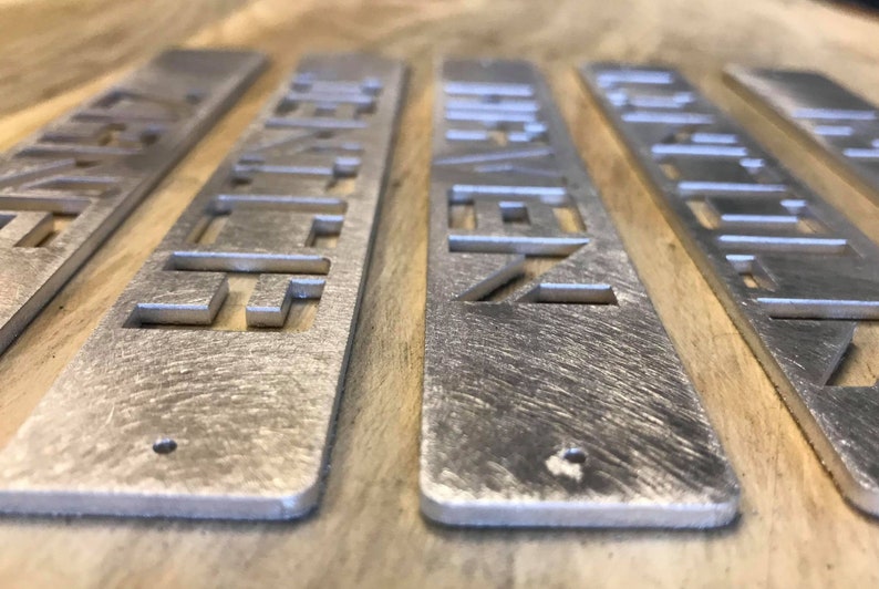 May include: Five silver metal rectangular signs with a geometric design cut out. The signs are arranged in a row on a wooden surface.