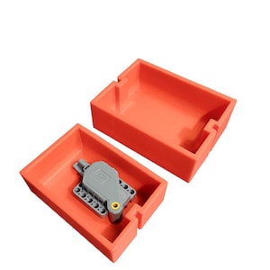 Könnte beinhalten: Offene orangefarbene rechteckige Box mit einer grauen Komponente im Inneren. Die Komponente hat einen gelben Kreis und mehrere kleine Löcher. Die Box ist offen, der Deckel leicht von der Basis getrennt, wodurch der Inhalt sichtbar wird.