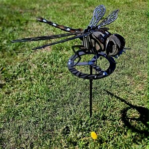 Pode incluir: Um ornamento de jardim em forma de lib&eacute;lula de metal escuro, com padr&otilde;es intrincados de asas recortadas e uma base circular. A escultura &eacute; constru&iacute;da com componentes de metal em camadas, exibindo uma cabe&ccedil;a, t&oacute;rax e abd&oacute;men detalhados numa haste vertical.