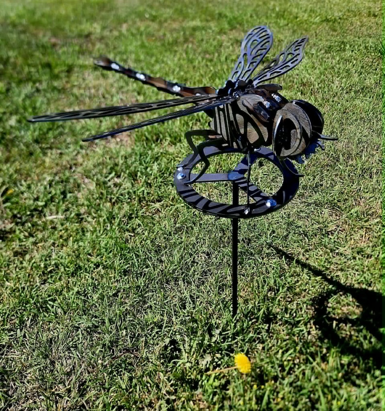 Pode incluir: Um ornamento de jardim em forma de lib&eacute;lula de metal escuro, com padr&otilde;es intrincados de asas recortadas e uma base circular. A escultura &eacute; constru&iacute;da com componentes de metal em camadas, exibindo uma cabe&ccedil;a, t&oacute;rax e abd&oacute;men detalhados numa haste vertical.