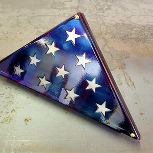 Könnte beinhalten: Ein dreieckiger, irisierender blau-violetter Flaggen-Display-Koffer mit weißen Sternaussparungen. Der Koffer hat eine glänzende Oberfläche und goldfarbene Eckakzente. Das Design erinnert an die amerikanische Flagge.