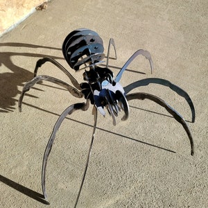 May include: A metal spider sculpture with a segmented head and body, and long, curved legs. The spider is made of dark metal and sits on a concrete surface. The sculpture casts a shadow.