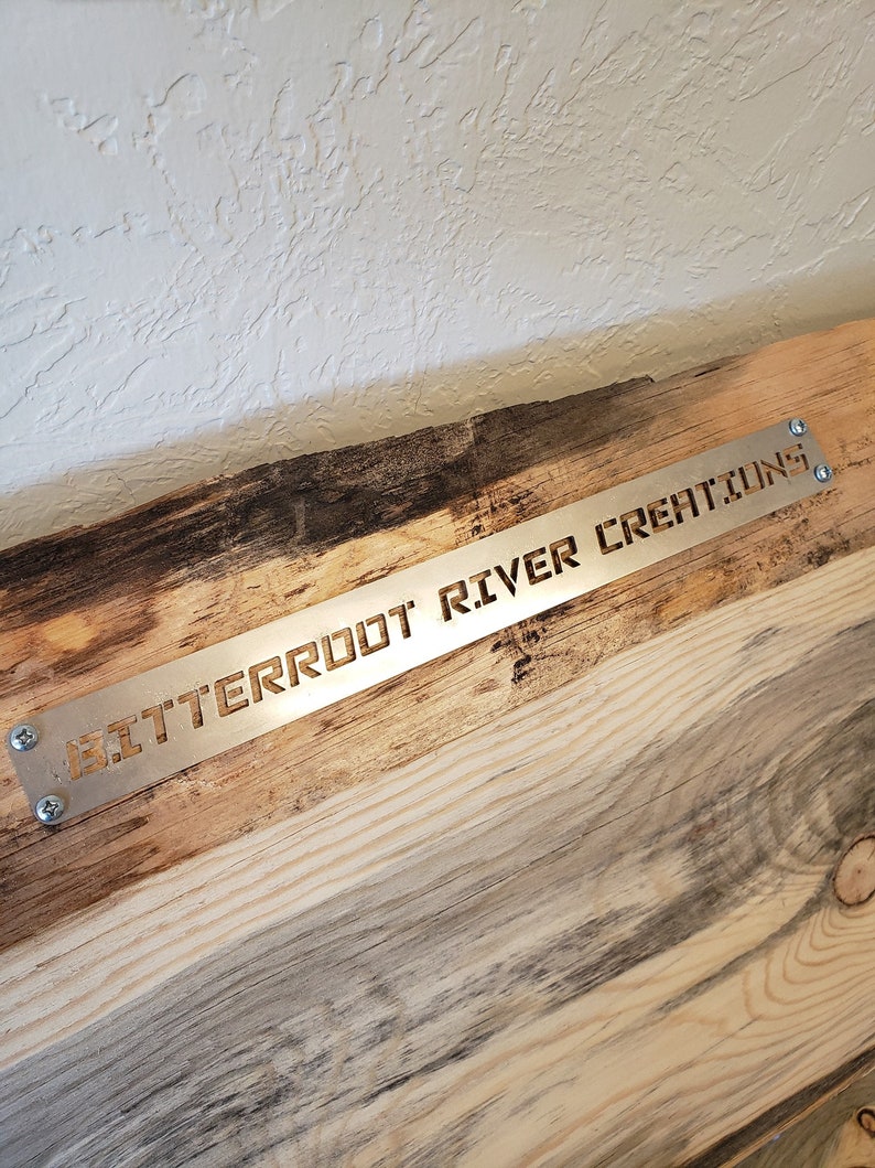 May include: A close-up of a wooden sign with a metal plate attached to it. The metal plate has the words "Bitterroot River Creations" cut out of it.