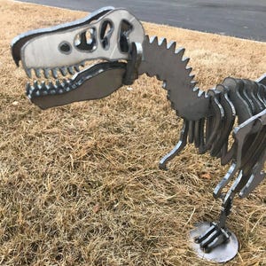 以下が含まれることがあります： 詳細な骨格デザインの金属製恐竜彫刻。 彫刻は、歯が見える大きな頭と、ギザギザのエッジを持つ背骨が特徴です。 恐竜は、銀色の金属で作られた円形の土台の上に立っています。