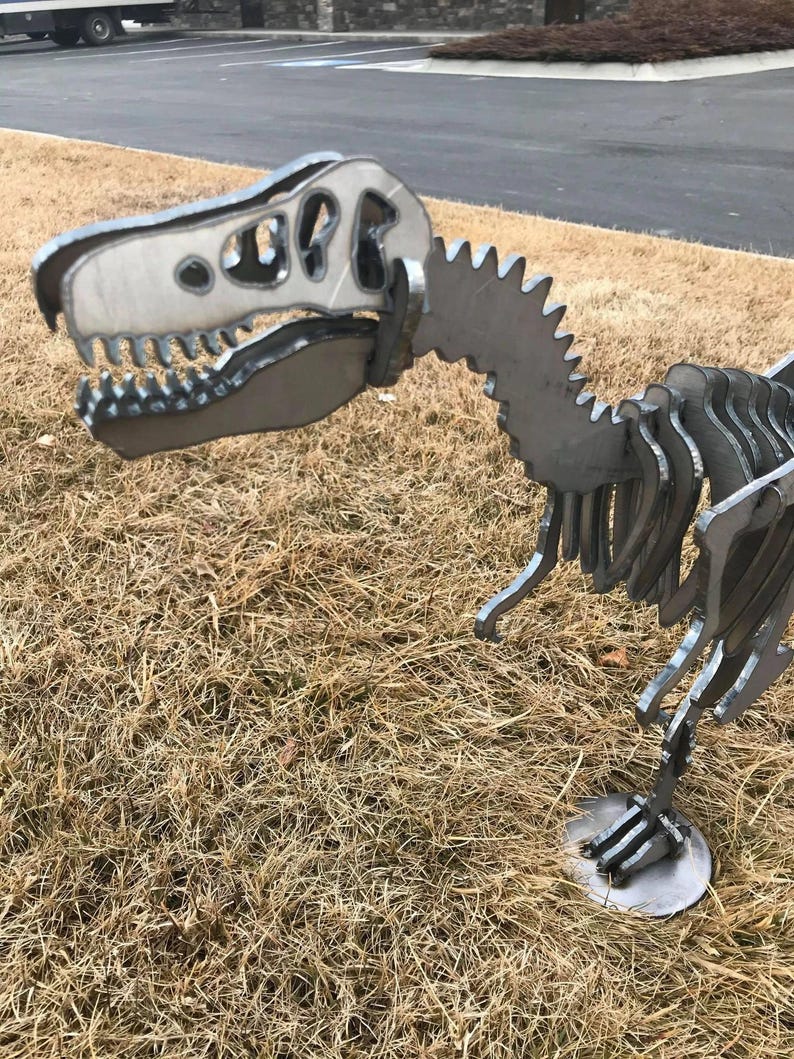 以下が含まれることがあります： 詳細な骨格デザインの金属製恐竜彫刻。 彫刻は、歯が見える大きな頭と、ギザギザのエッジを持つ背骨が特徴です。 恐竜は、銀色の金属で作られた円形の土台の上に立っています。