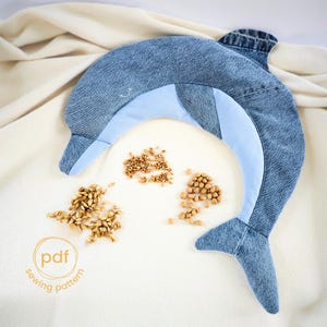 Puede incluir: Un animal de peluche de delfín de mezclilla azul con vientre azul claro. El delfín está acostado sobre una superficie blanca con un círculo que dice "pdf sewing pattern" y un montón de pequeñas semillas marrones.
