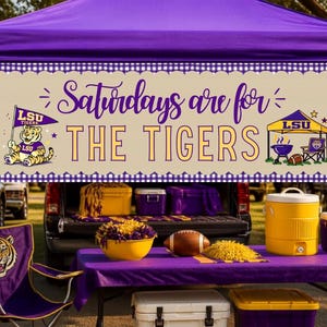 Puede incluir: Una pancarta rectangular con fondo beige y borde de cuadros morados que incluye el texto "Saturdays are for THE TIGERS". El diseño muestra un tigre de dibujos animados con una bandera de LSU, una tienda morada y dorada, una parrilla y un balón.