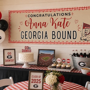 May include: A rectangular banner with a red and white gingham border, beige centre, and text reading CONGRATULATIONS Anna Kate GEORGIA BOUND. It features a Georgia Bulldogs mascot illustration, red hearts, and a black table with party decor.