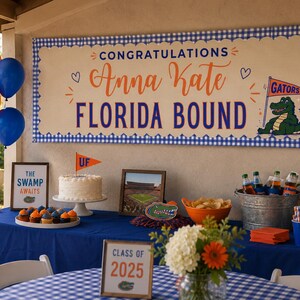 May include: A large rectangular banner with a blue and white gingham border, featuring the text CONGRATULATIONS Anna Kate FLORIDA BOUND, a small orange pennant with GATORS, and a green alligator illustration on a cream-coloured background.