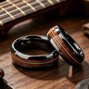 May include: Two rings with a polished black finish, featuring a central inlay of brown wood grain and a thin, metallic guitar string wrapped around the circumference. The rings have a smooth, reflective surface and a rounded, domed profile.