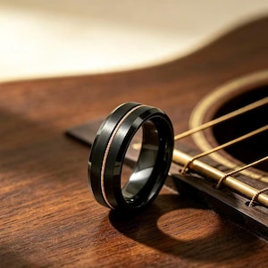 May include: A black ring with a brushed finish and a thin, copper-colored wire inlay running horizontally around the center. The band features beveled edges and a smooth, polished interior surface, showcasing a distinct metallic contrast.