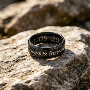Op de afbeelding: Een zwarte ring met de inscriptie "1-09-20" en "always & forever" in witte letters. De ring is geplaatst tegen een getextureerde, lichtgekleurde stenen achtergrond. De ring lijkt van metaal te zijn.