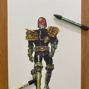 Peut inclure: Illustration de Judge Dredd en uniforme complet, avec une combinaison noire et verte, des épaulettes dorées et un casque à visière rouge. L'œuvre est accompagnée de deux stylos ProMarker noirs sur une surface en bois.