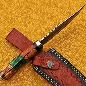 以下が含まれることがあります： 木製のハンドルと革製の鞘が付いた狩猟用ナイフ。ハンドルは緑と茶色のアクセントが付いた木で作られています。ブレードは鋼製で、ダマスカス模様が施されています。鞘は茶色の革製で、装飾的なステッチが施されています。