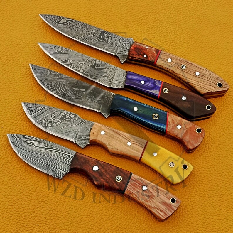 May include: Five Damascus steel hunting knives with wooden handles. The handles are made of different types of wood and have different colors, including red, purple, blue, yellow, and brown.