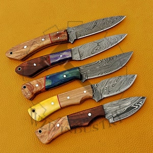 May include: Five Damascus steel knives with wooden handles. The handles are made of different types of wood, including a light wood with a yellow accent, a dark wood with a red accent, and a blue and black wood with a red accent.