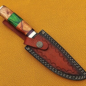 以下が含まれることがあります： 装飾的なステッチパターンが施された茶色の革製のナイフシース。シースには金属製のボタンが付いており、緑と茶色の木製のハンドルが付いたナイフに取り付けられています。