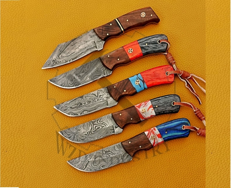 May include: Set of five Damascus steel knives with wooden handles and colorful inlays. Each knife has a different colored inlay, including red, blue, and black. The knives are arranged in a row on a yellow surface.