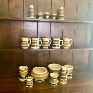 May include: A collection of vintage ceramic tableware with a cream base, featuring horizontal bands of brown, green, and orange. The set includes jugs, mugs, bowls, and decorative vases, arranged on wooden shelves.