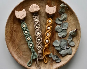 May include: Three wooden baby teethers with fox, rainbow, and fox designs. Each teether is attached to a macrame cord with wooden beads spelling out the names Liam, Elise, and Liam.