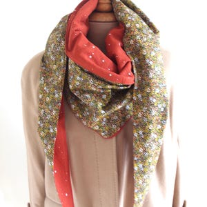 Peut inclure: Une écharpe réversible avec un motif floral d'un côté et une couleur rouge unie avec des pois blancs de l'autre. L'écharpe est présentée sur un mannequin portant un manteau marron.