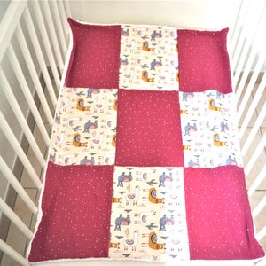Peut inclure: Une couverture bébé en patchwork avec une bordure blanche. La couverture est faite de quatre tissus différents, chacun avec un motif différent. Un tissu a un fond rose avec des pois blancs, un autre a un fond crème avec des lamas et des cactus bruns, et les deux autres ont un fond crème avec des lamas et des cactus gris.