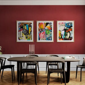 May include: Three framed abstract art pieces with colorful, expressive designs hang above a dark wood dining table. The room features a deep red wall, black chairs, and a modern aesthetic. A plant in a pot and a candle holder are also visible.