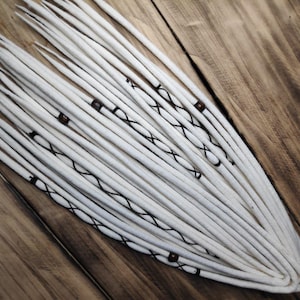 Könnte beinhalten: Weiße synthetische Dreadlocks mit schwarzen Akzenten und braunen Perlen. Die Dreadlocks sind in einem Bündel angeordnet und liegen auf einer Holzoberfläche.