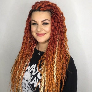 May include: A woman with long, thick braids in shades of orange, red, and blonde. She is wearing a black shirt with a white graphic print that says "WAG" and "LAW".