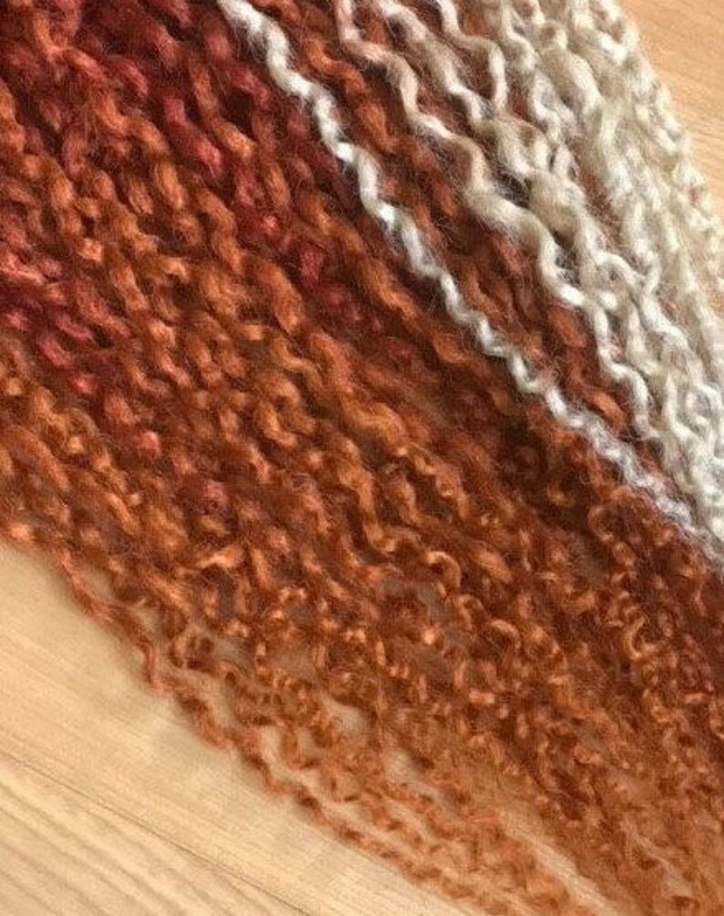 May include: A bundle of curly hair extensions in shades of brown, orange, and white. The hair is arranged in a gradient from dark brown to light blonde.