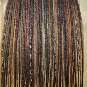May include: A collection of handmade dreadlocks in various shades of brown, auburn, and olive green. The dreads are arranged in vertical rows, with white thread accents. The overall look is natural and textured, suitable for hair styling.