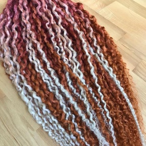 May include: A bundle of long, curly hair extensions in shades of red, orange, and white. The hair is arranged in rows, with the red shades at the bottom and the white shades at the top.