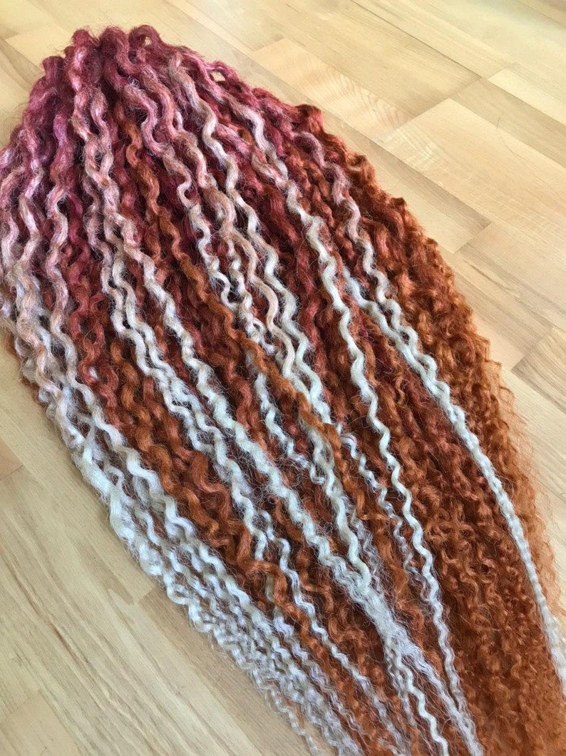 May include: A bundle of long, curly hair extensions in shades of red, orange, and white. The hair is arranged in rows, with the red shades at the bottom and the white shades at the top.