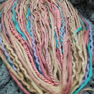 May include: A collection of handmade dreadlocks in various shades of pink, blue, and beige. Some dreads are braided or adorned with beads and small silver accents. The bundle is tied together with twine, creating a bohemian aesthetic.