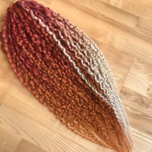 May include: A long strand of ombre hair extensions with a red, orange, and blonde color gradient. The hair is in a curly style.