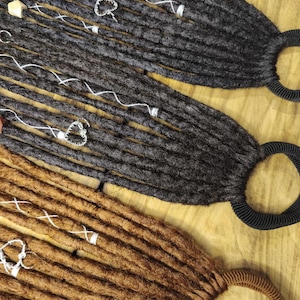 Può includere: Primo piano di tre extension per dreadlock nei toni del marrone e del grigio. Ogni dreadlock è adornato con filo bianco e perline decorative. Le extension sono attaccate a elastici neri o marroni.