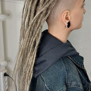 May include: A person with a shaved head and long, light blonde dreadlocks. They are wearing a black hooded sweatshirt and a blue denim jacket.